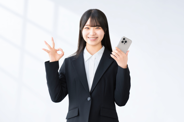 スマホを片手にOKのポーズをするスーツの女性