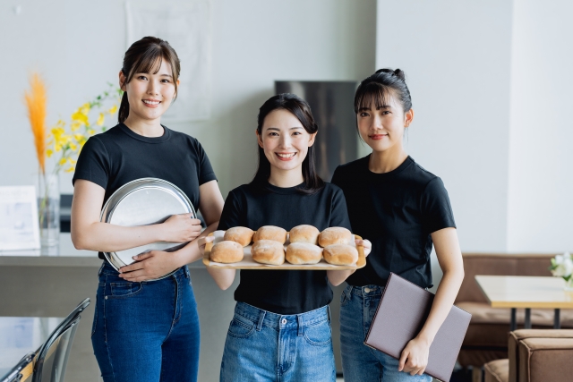 それぞれトレーや商品、メニュー表を持ってこちらに微笑む飲食店の3人の女性スタッフ