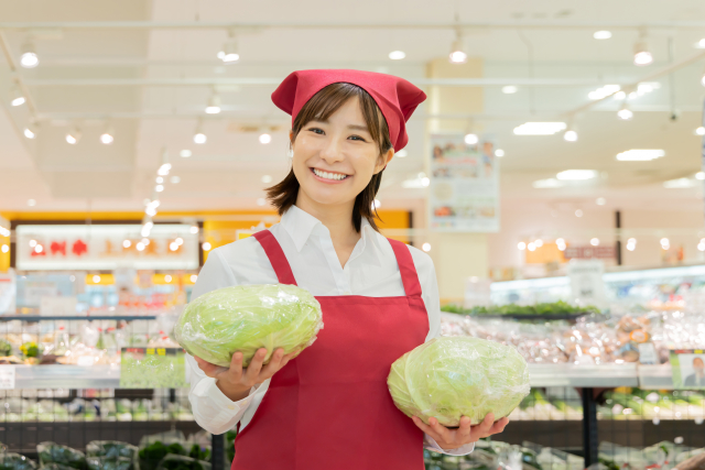 両手に野菜を持ちながらこちらに微笑むスーパーの女性店員