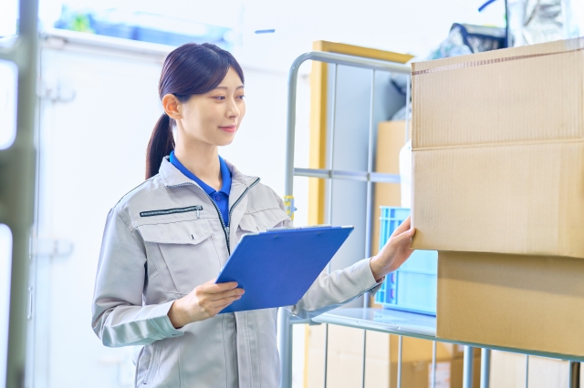倉庫内でバインダーを手に商品のチェックを行うポニーテールの女性スタッフ