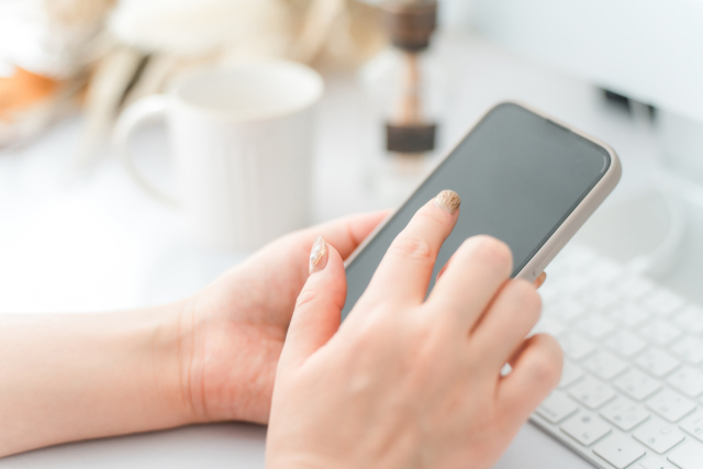 ベージュのネイルをした女性の手がスマートフォンを操作しているイメージ