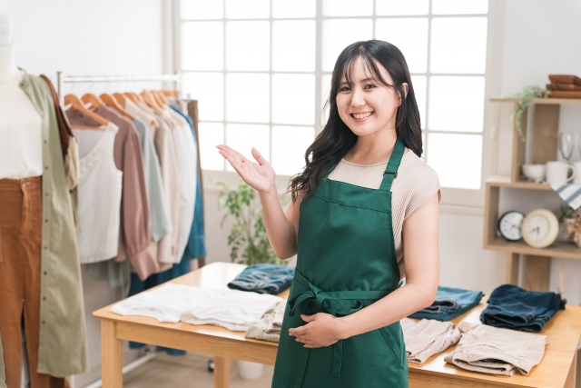 服が並べられた店内を指し示しながらこちらに微笑むアパレル店の女性スタッフ