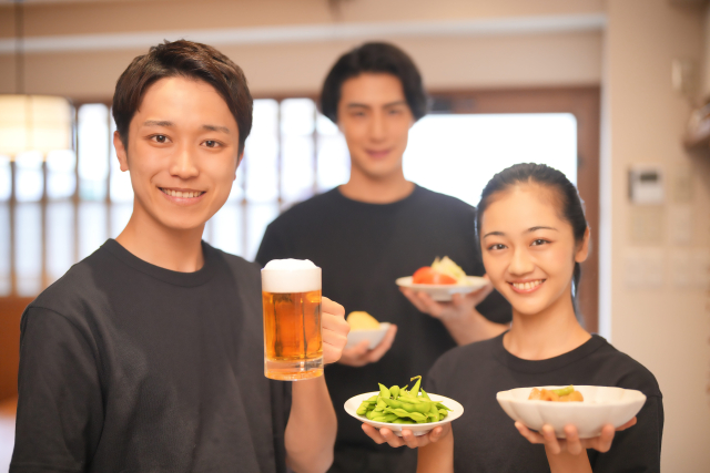 ビールと枝豆とおかずを持って微笑む男女3人のアルバイトスタッフ
