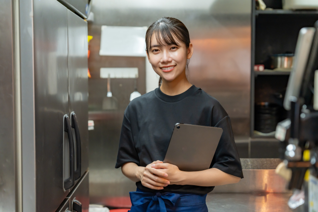 飲食店のバックルームでタブレットを抱えながらこちらに微笑む女性スタッフ