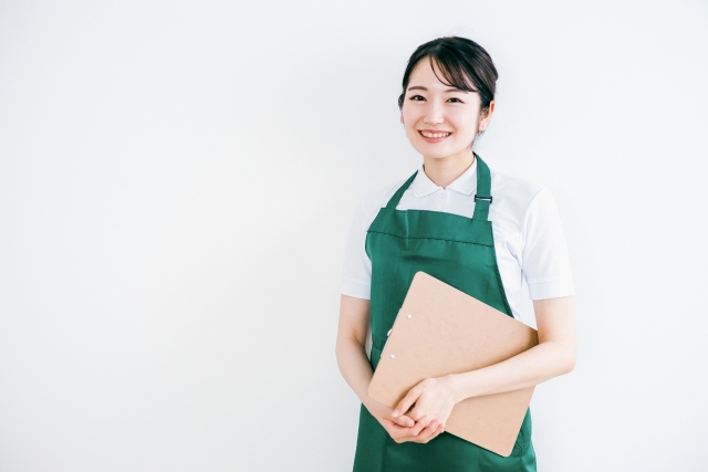 バインダーを抱えながらこちらに微笑む緑のエプロン姿の女性スタッフ