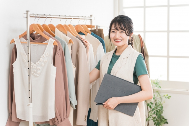 在庫管理用タブレットをわきに抱えてこちらに振り向くアパレル店の女性スタッフ