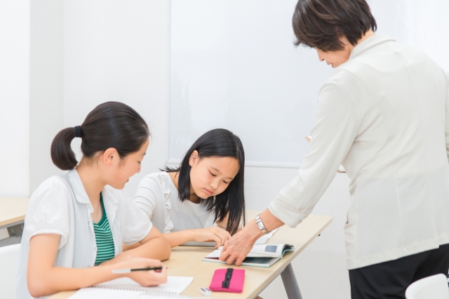 机に並んで座る女子生徒2名に勉強を教える女性塾講師