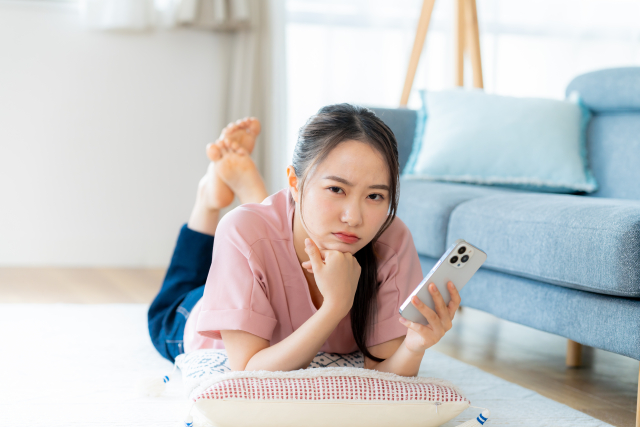 自室でスマホを見ながら困った顔をする女性